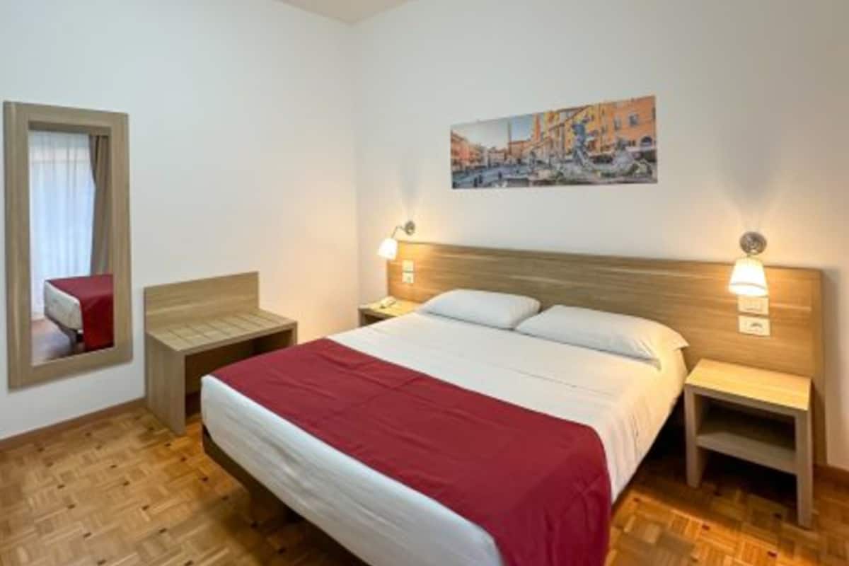 A hotel room with a wooden floor, a bed with white and red bedding, two bedside tables with lamps, a painting on the wall, and a window with curtains.