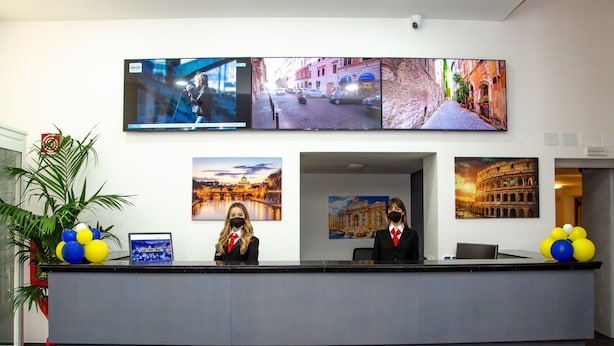 A reception desk with a receptionist, a large flat-screen TV displaying cityscapes, and framed pictures of famous landmarks.