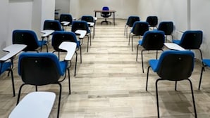 A conference room with rows of blue and black chairs, white tables, and a central table with a blue chair. The room has a tiled floor and white walls with a window on the left.