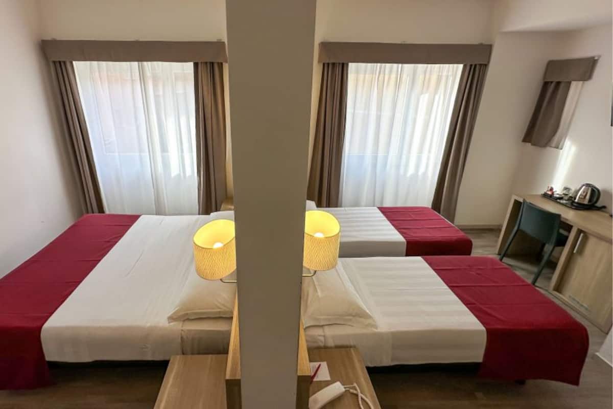 A hotel room with two beds, each with a red and white bedspread, and a bedside lamp. There is a desk with a chair and a kettle on it.