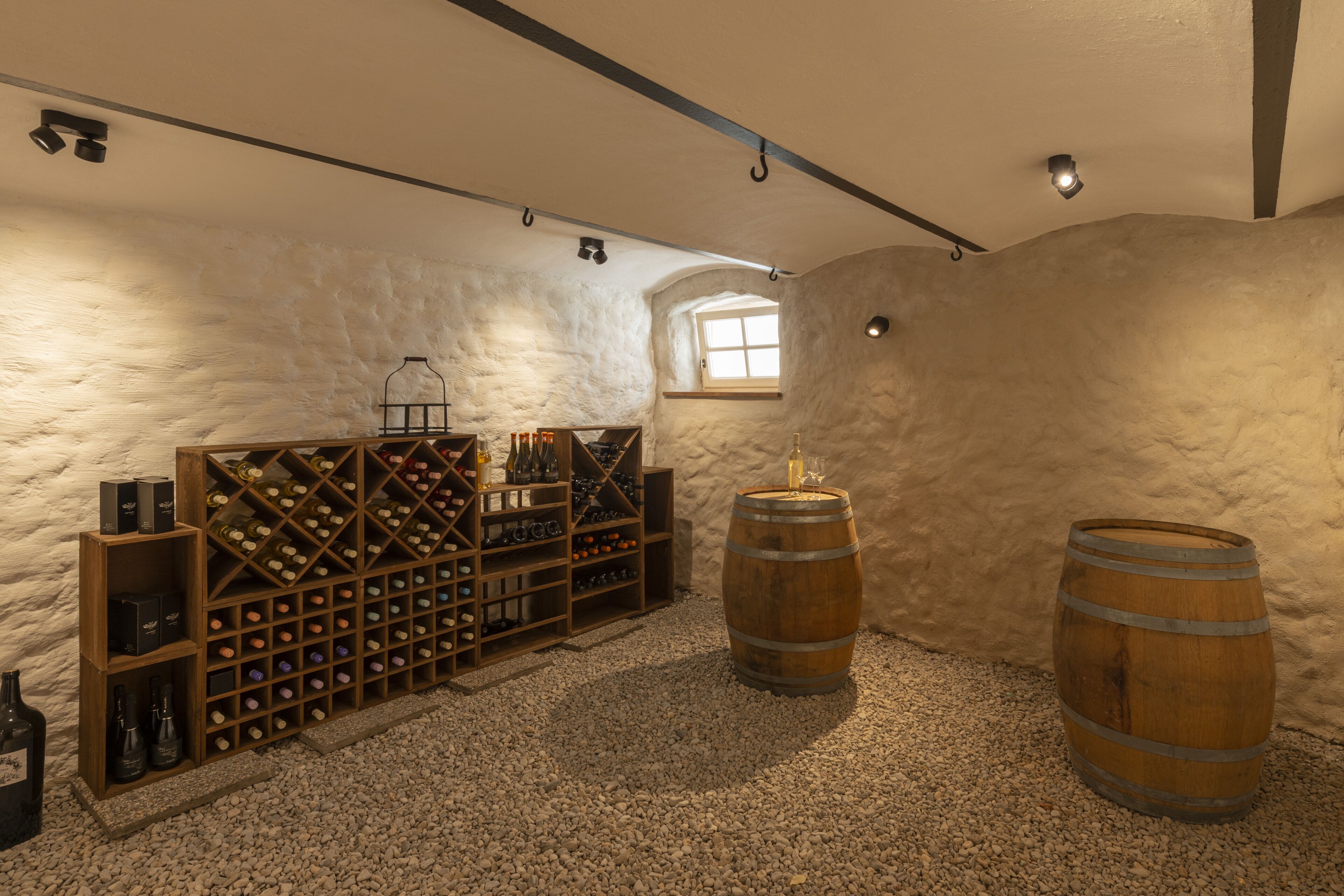 Une cave à vin avec des étagères en bois pour les bouteilles, deux barriques et une petite fenêtre.
