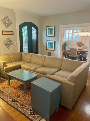 A living room with a sectional sofa, two side tables, a patterned rug, and a dining area with a chandelier.