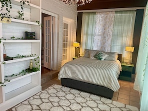 A bedroom with a bed, two bedside tables with lamps, a white brick wall, a door, and shelves with plants.