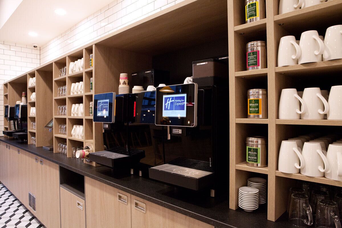A hotel reception area with a self-service coffee station, shelves with various items, and a counter with a digital display.