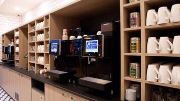 A hotel reception area with a self-service coffee station, shelves with various items, and a counter with a digital display.