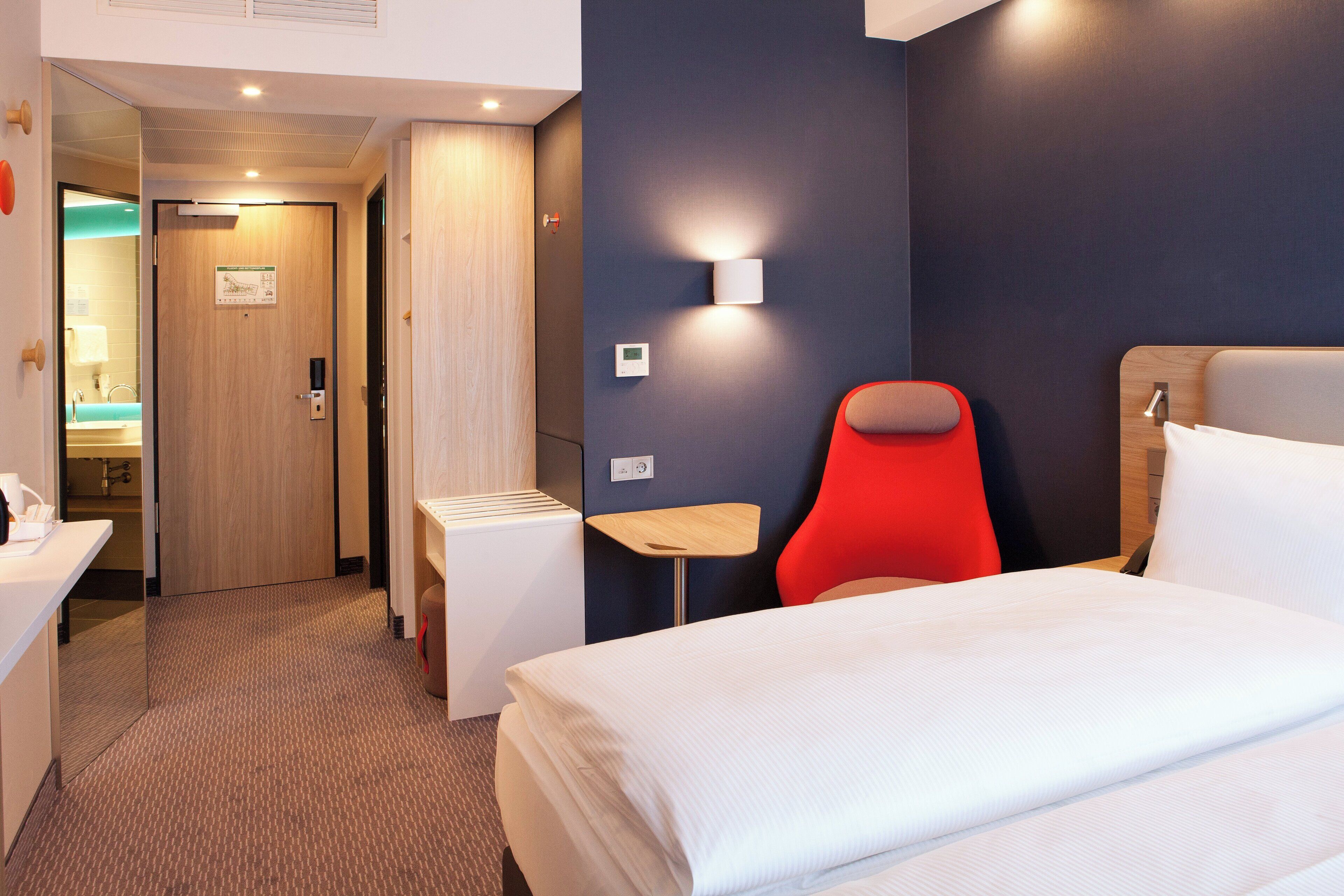 Een hotelkamer met een bed, een rode stoel, een klein tafeltje, een wandlamp en een deur naar een badkamer.