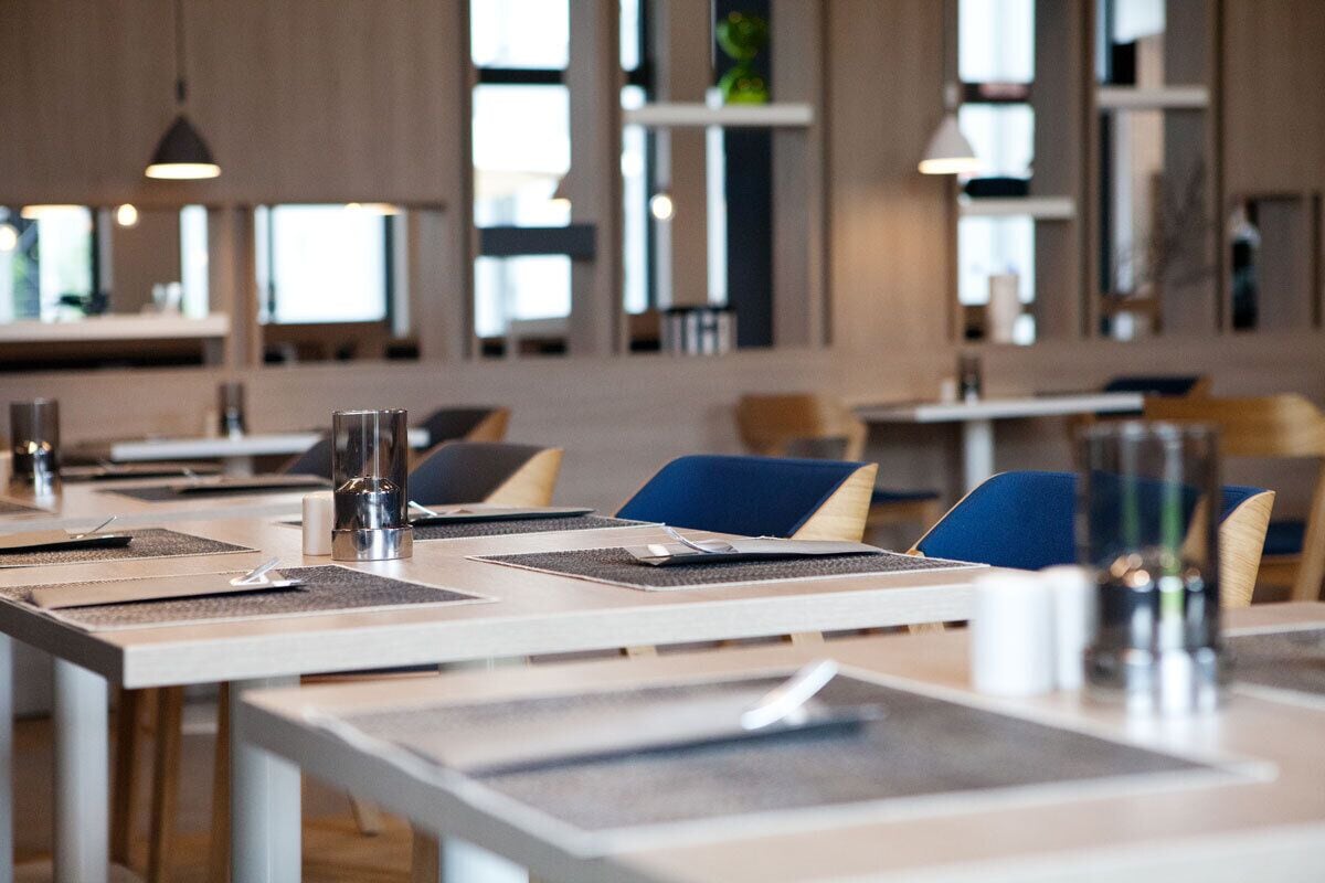 Un espace de restauration moderne avec des tables prévues pour deux personnes, caractérisé par un décor minimaliste et une lumière naturelle abondante grâce à de grandes fenêtres.