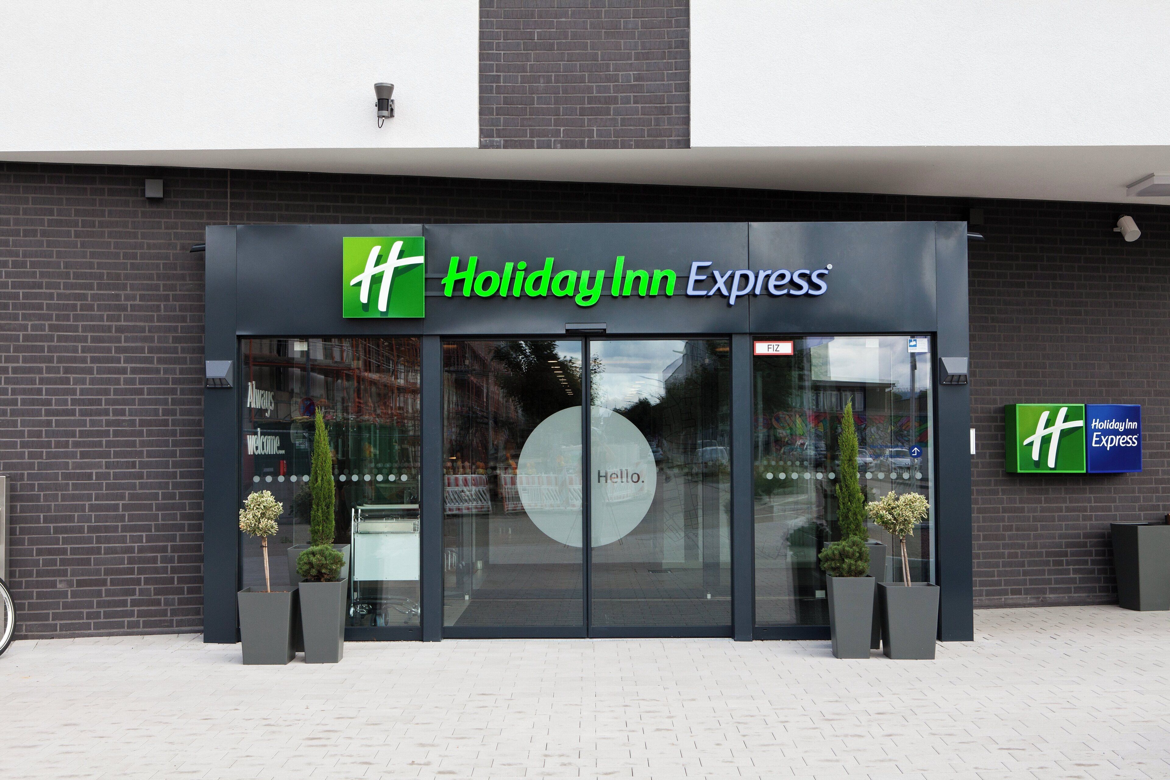 Entrada de un hotel Holiday Inn Express con puerta de vidrio, un letrero que dice 'Hello' y dos plantas en macetas.