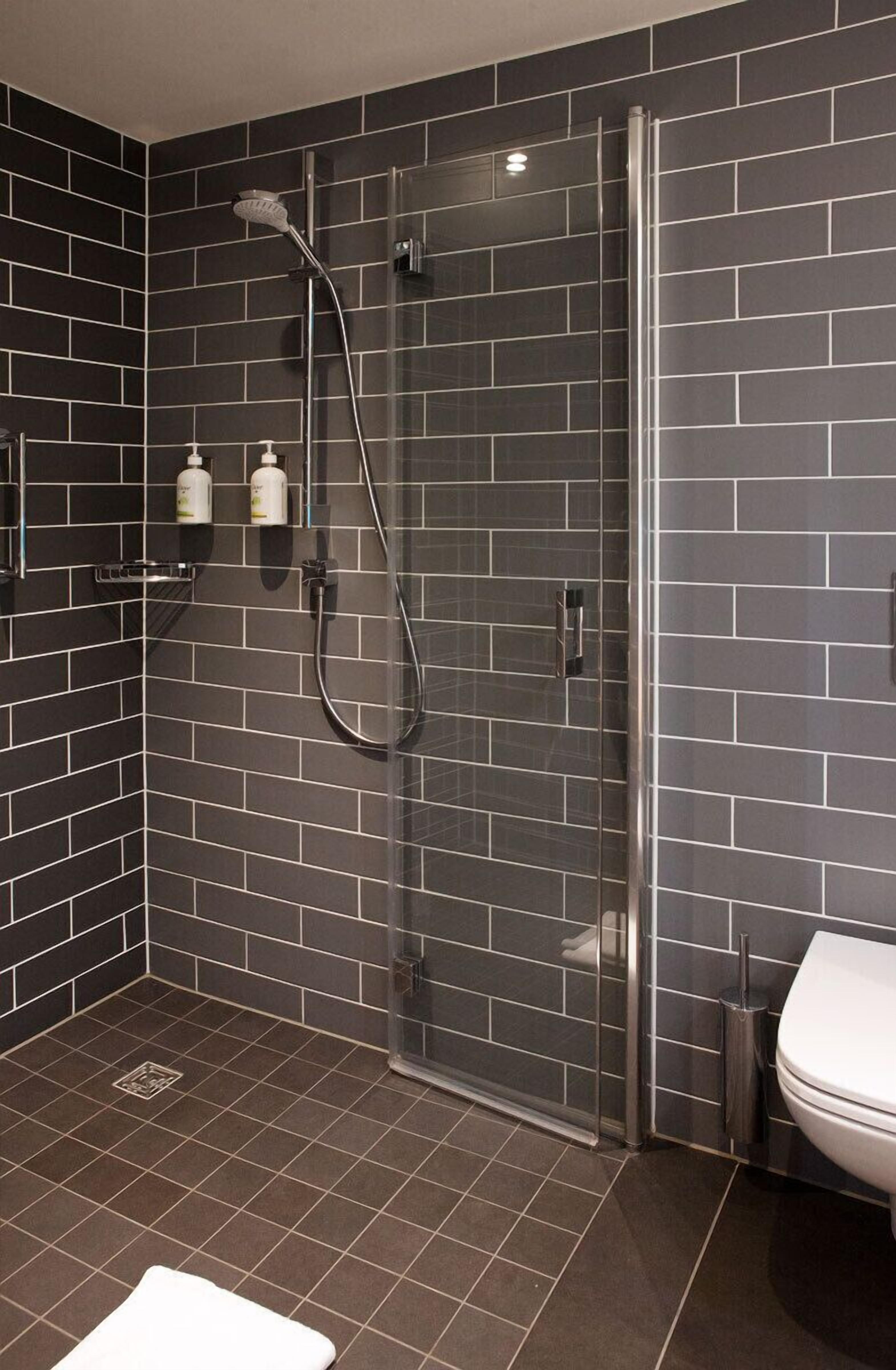 Un baño moderno con paredes de azulejos oscuros, un cerramiento de ducha de vidrio, inodoro blanco y un pequeño estante con artículos de aseo.