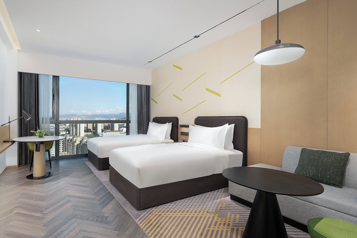 Habitación de hotel con dos camas, una mesa de comedor, un sofá, vista a la ciudad y un gran ventanal.