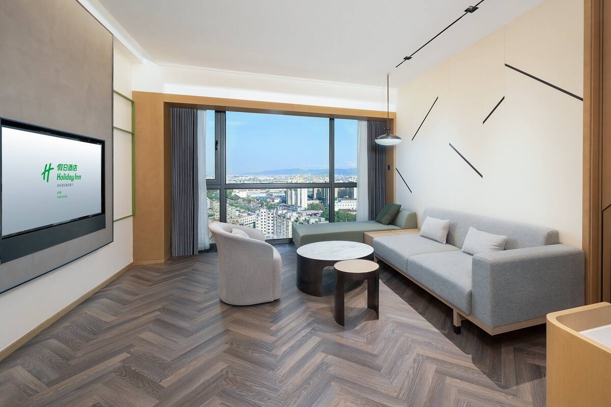 A modern hotel room with a large window offering a city view, a sofa, a chair, and a small table.