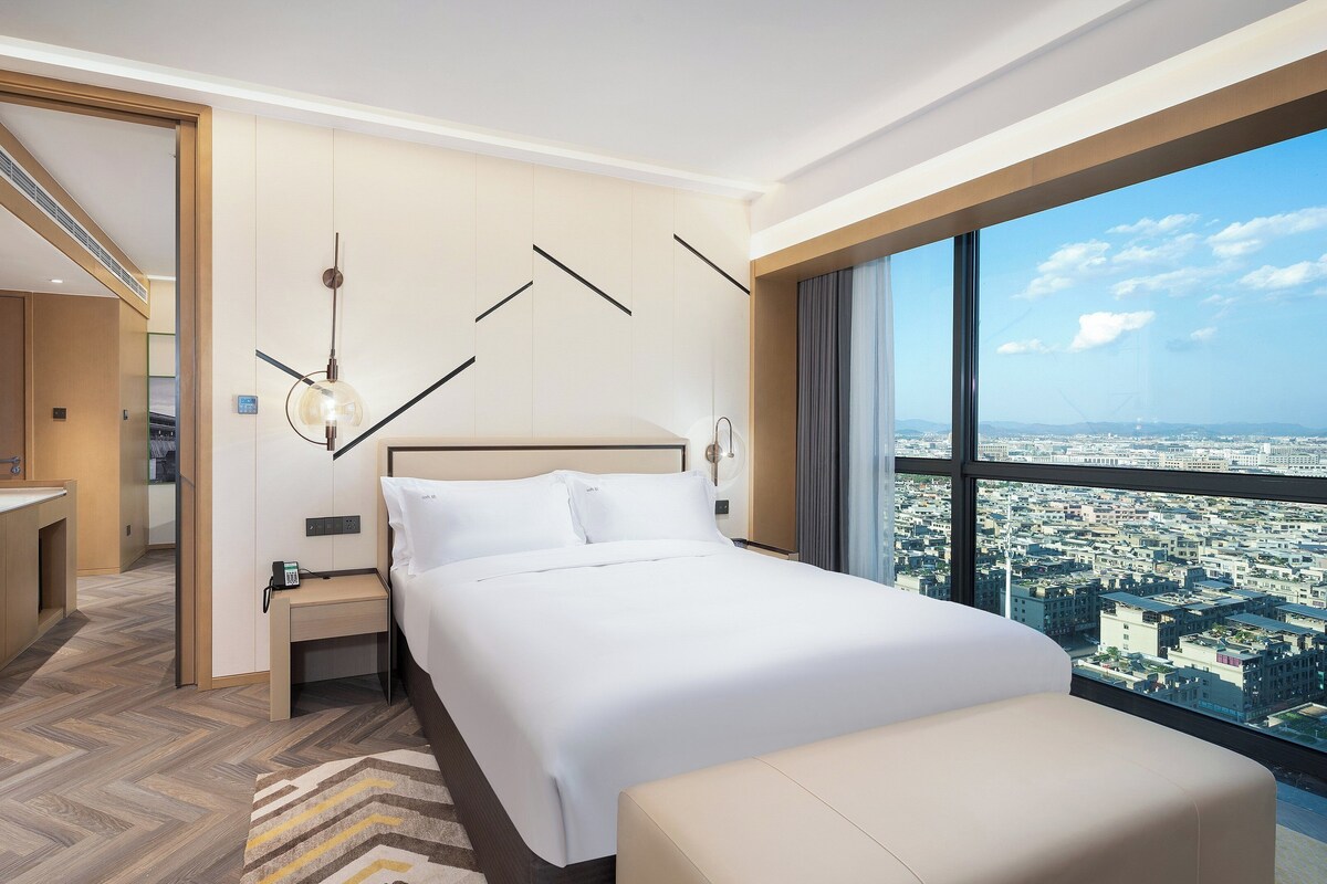 Habitación de hotel con una cama grande, una mesita de noche con teléfono, una lámpara moderna y vistas a la ciudad a través de amplios ventanales.