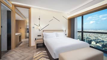 A hotel room with a large bed, a bedside table with a phone, a modern light fixture, and a city view through large windows.