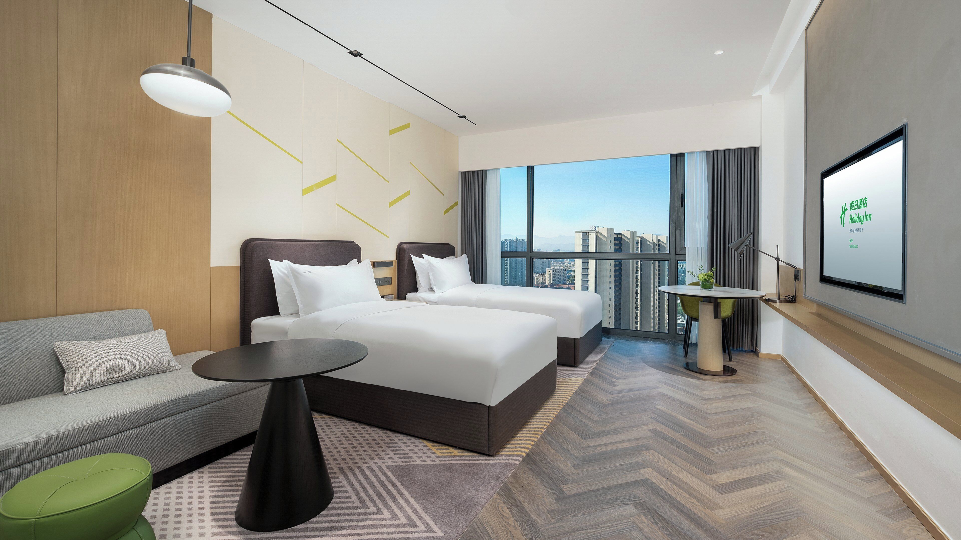 A modern hotel room with a large bed, a sofa, a small table, and a view of the city through large windows.