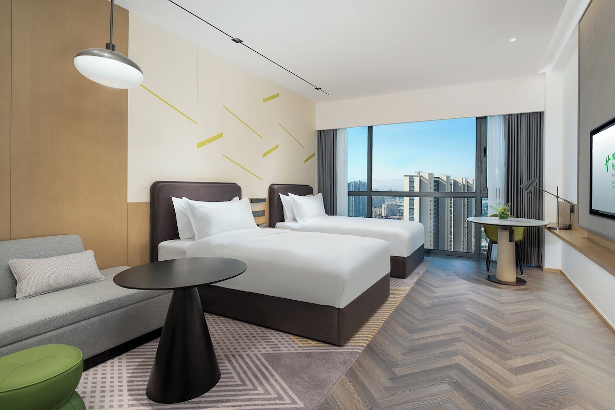 Habitación de hotel moderna con una cama grande, un sofá, una mesita y vistas de la ciudad a través de amplios ventanales.