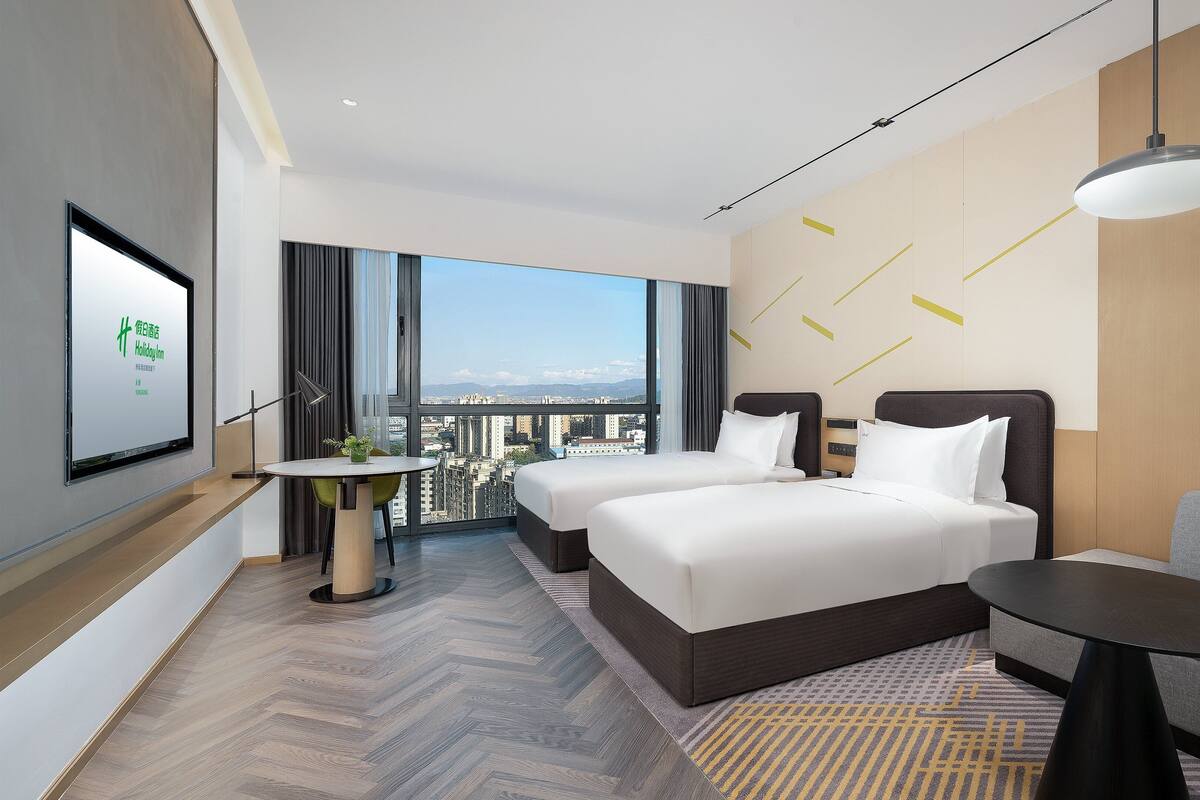 A modern hotel room with two beds, a balcony with a city view, a small table, and a sofa.