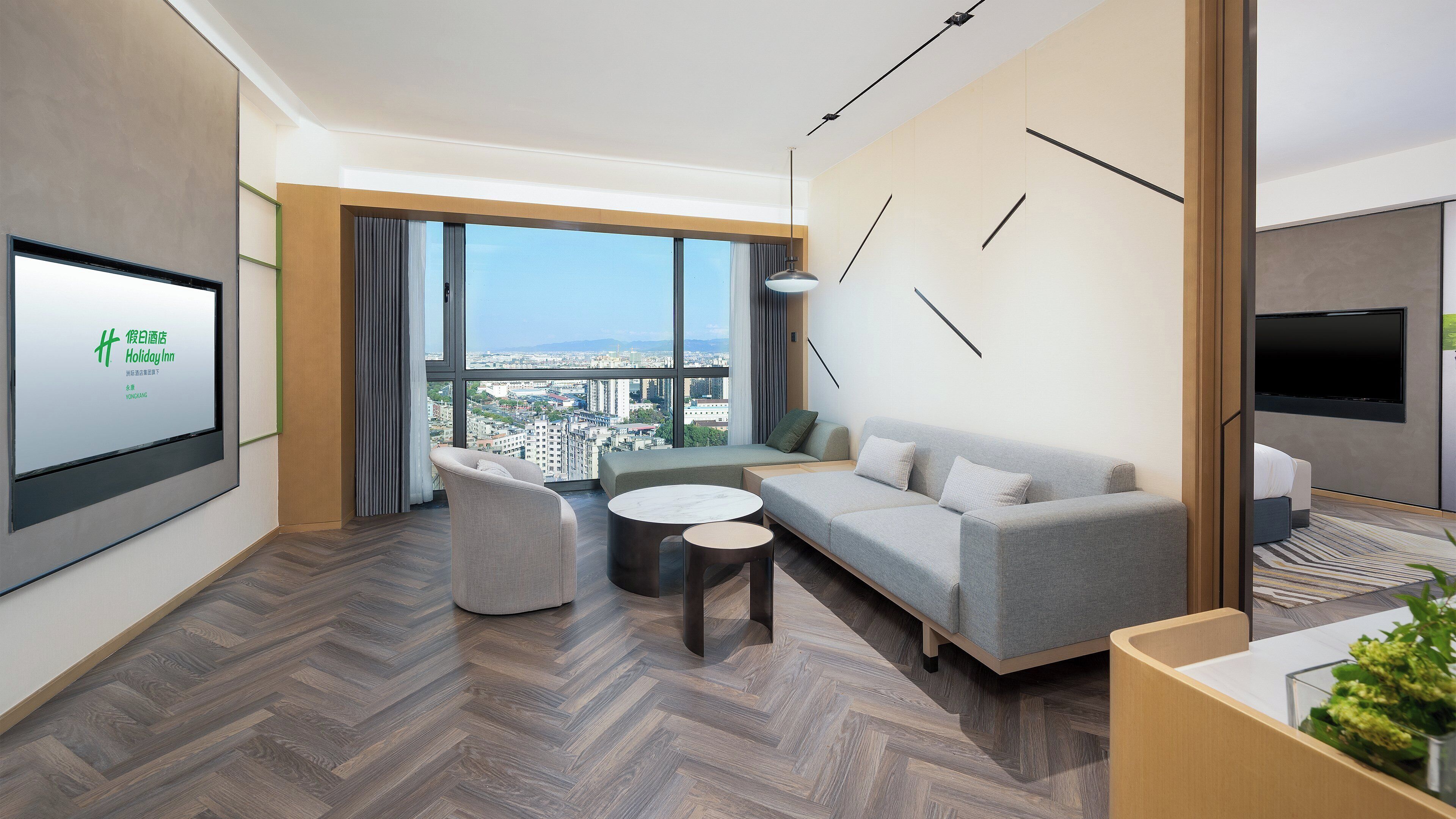 A modern hotel room with a large window offering a city view, a sofa, a chair, and a small table.