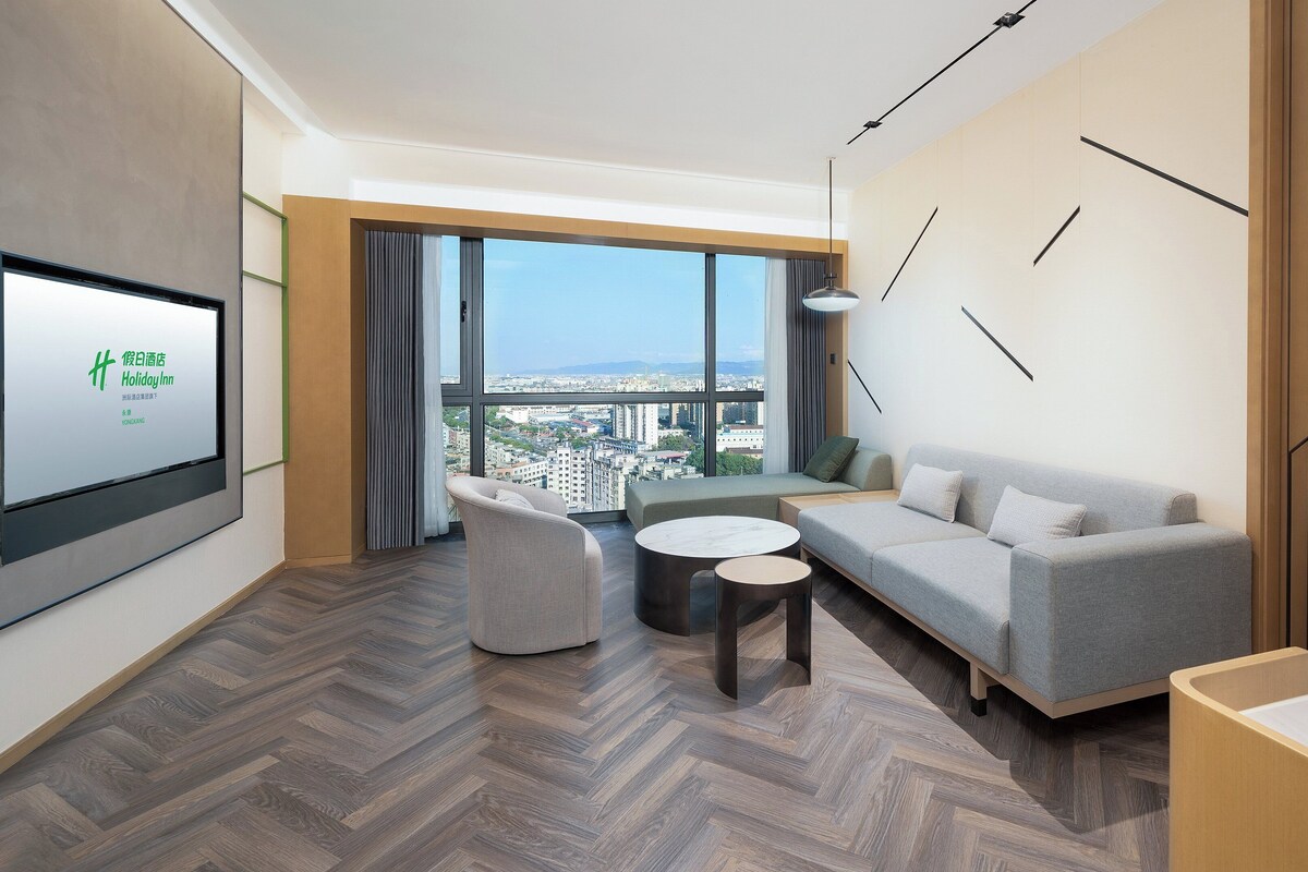 Una habitación de hotel moderna con un ventanal grande que ofrece vistas a la ciudad, un sofá, una silla y una mesita.
