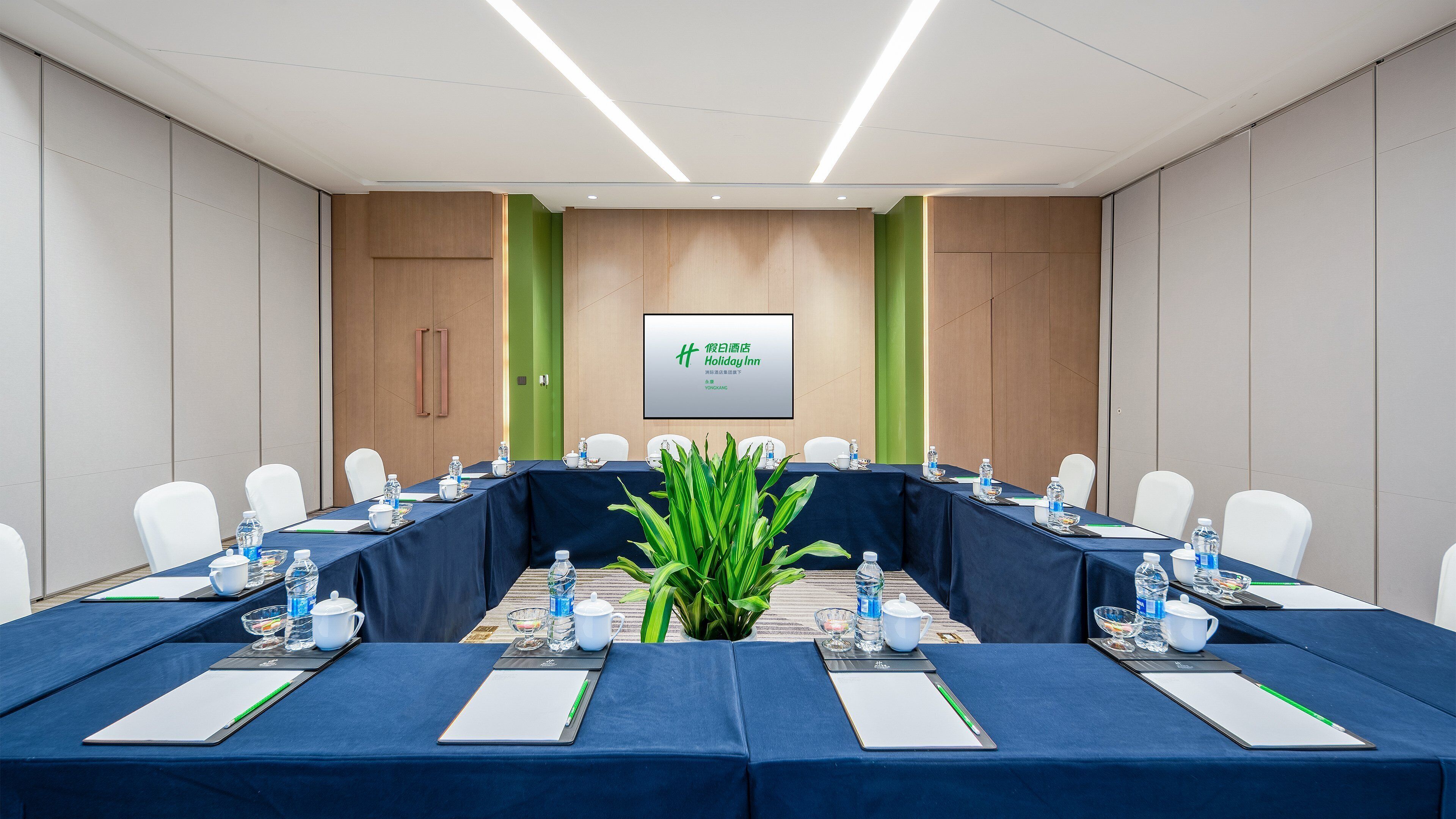 Un salón de conferencias con una larga mesa, sillas blancas y una pantalla grande que muestra el logotipo de Holiday Inn.