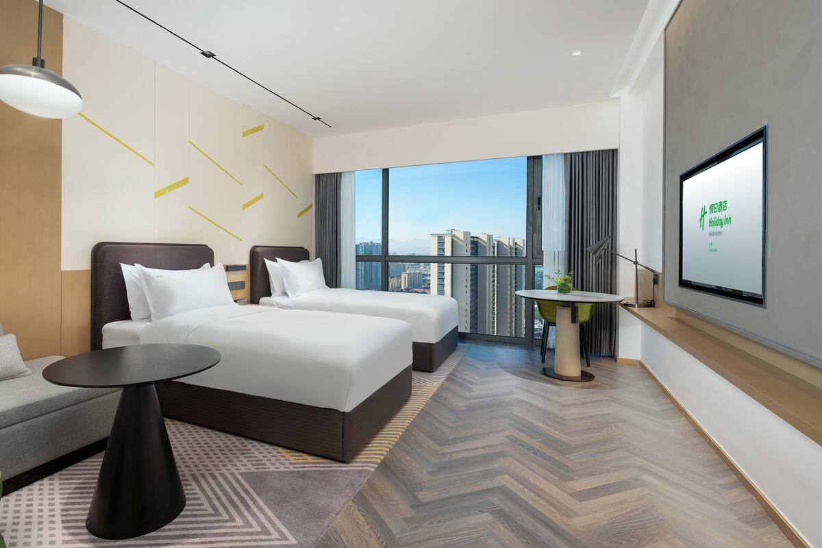 A modern hotel room with a large bed, a sofa, a small table, and a view of the city through large windows.