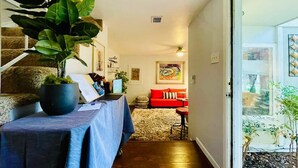 Una sala de estar con una escalera, un sofá rojo, una mesa con mantel azul y una planta grande.