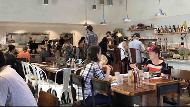 Un restaurante espacioso con techos altos, vigas a la vista e iluminación colgante. Las mesas están dispuestas para comer, y se ve la zona de la cocina al fondo.