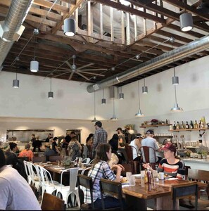 Un restaurante espacioso con techos altos, vigas a la vista e iluminación colgante. Las mesas están dispuestas para comer, y se ve la zona de la cocina al fondo.