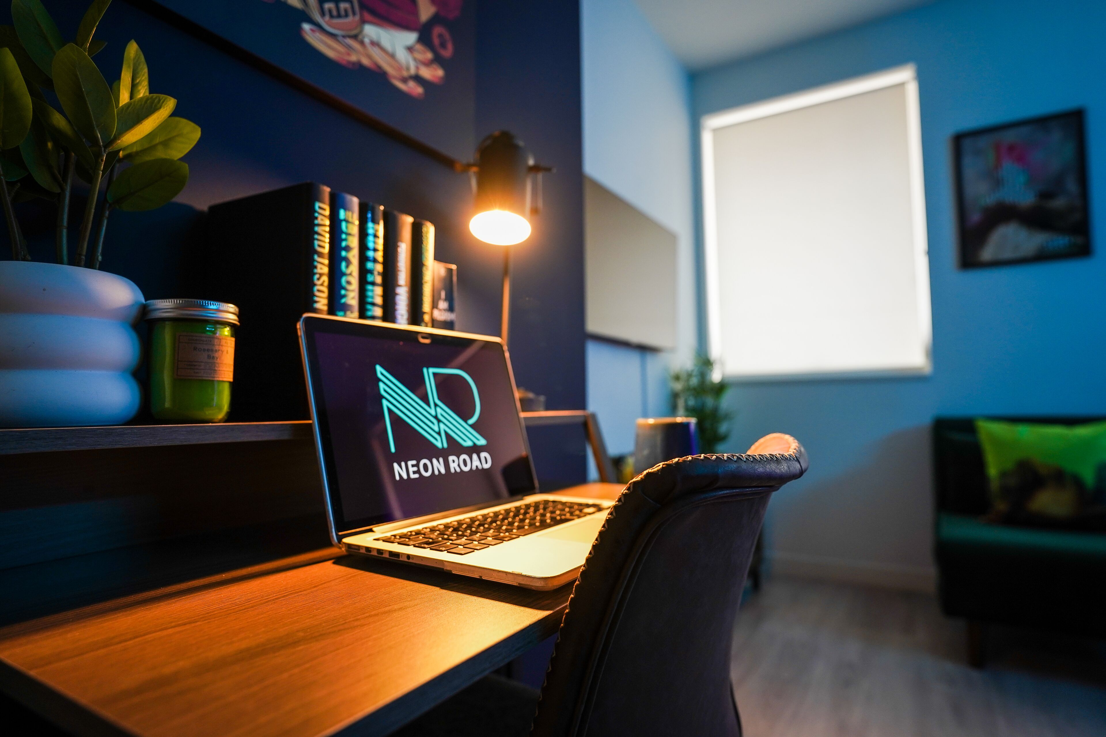 A laptop on a desk with a neon road sign, a plant, and a lamp.