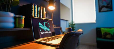 A laptop on a desk with a neon road sign, a plant, and a lamp.