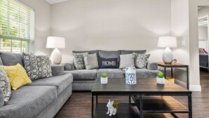 Una sala de estar moderna con un sofá seccional gris, dos mesitas de noche con lámparas, una mesa de centro y cojines decorativos.