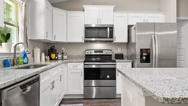 Una cocina moderna con armarios blancos, electrodomésticos de acero inoxidable, encimeras de granito y una ventana con una planta.