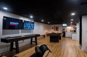 A modern living room with a pool table, a foosball table, and a billiards table. There are two large flat-screen TVs mounted on the wall.