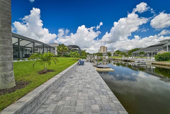A waterfront walkway with a clear sky, a calm water body, and modern buildings in the background.
