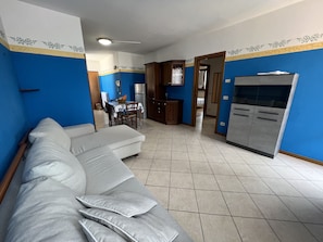 A living room with a sofa, a dining table with chairs, a kitchen area with cabinets and a refrigerator, and a hallway leading to another room.