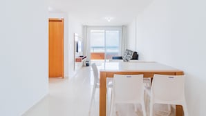 A modern, bright room with a wooden dining table and white chairs, a large window with a view of the sea, and a wooden door leading to another area. - PFL01 - Apartamento 2 Quartos -Vista Mar (Salvador)