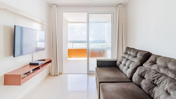A modern living room with a flat-screen TV, a wooden console table, a grey sofa, and a sliding glass door leading to a balcony with a view of the sea. - PFL01 - Apartamento 2 Quartos -Vista Mar (Salvador)
