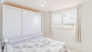 A bedroom with a bed, a wardrobe, a window with curtains, and a view of the beach. - PFL01 - Apartamento 2 Quartos -Vista Mar (Salvador)