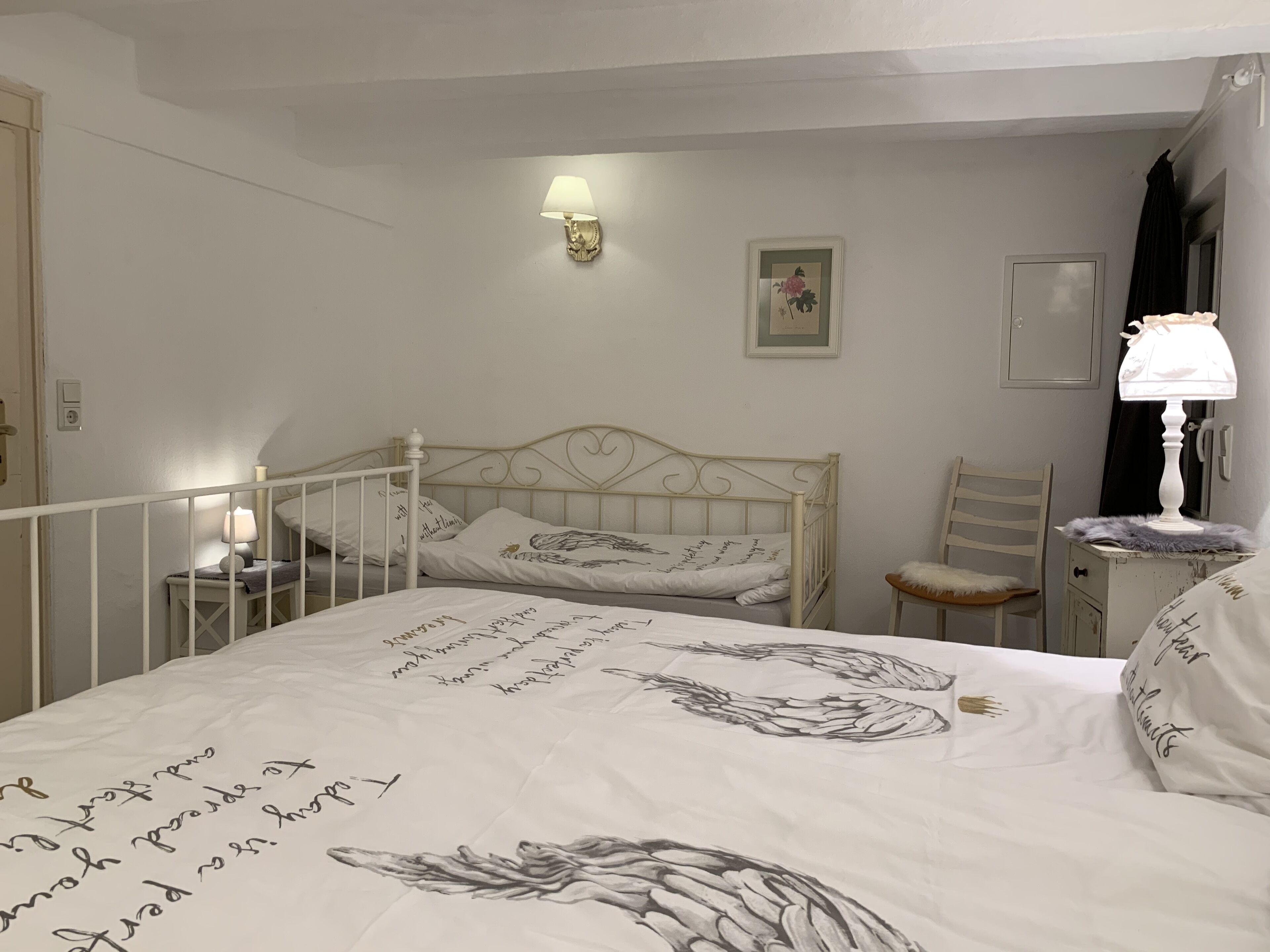 A bedroom with a bed, a chair, a nightstand, a wall-mounted lamp, and a framed picture.