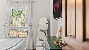 Un dormitorio con cama, espejo, escritorio con lámpara, ventana con cortinas y un letrero en la pared.