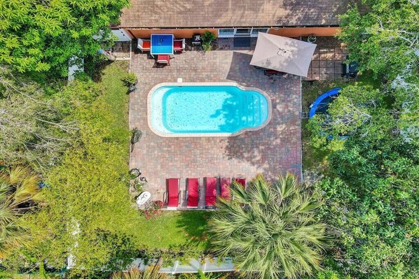 An aerial view of a swimming pool surrounded by a brick patio, with red lounge chairs and a small table.