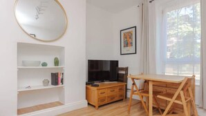 Une pièce avec un plancher en bois, un téléviseur posé sur un meuble en bois, une table à manger avec des chaises, un miroir au mur et un tableau encadré.