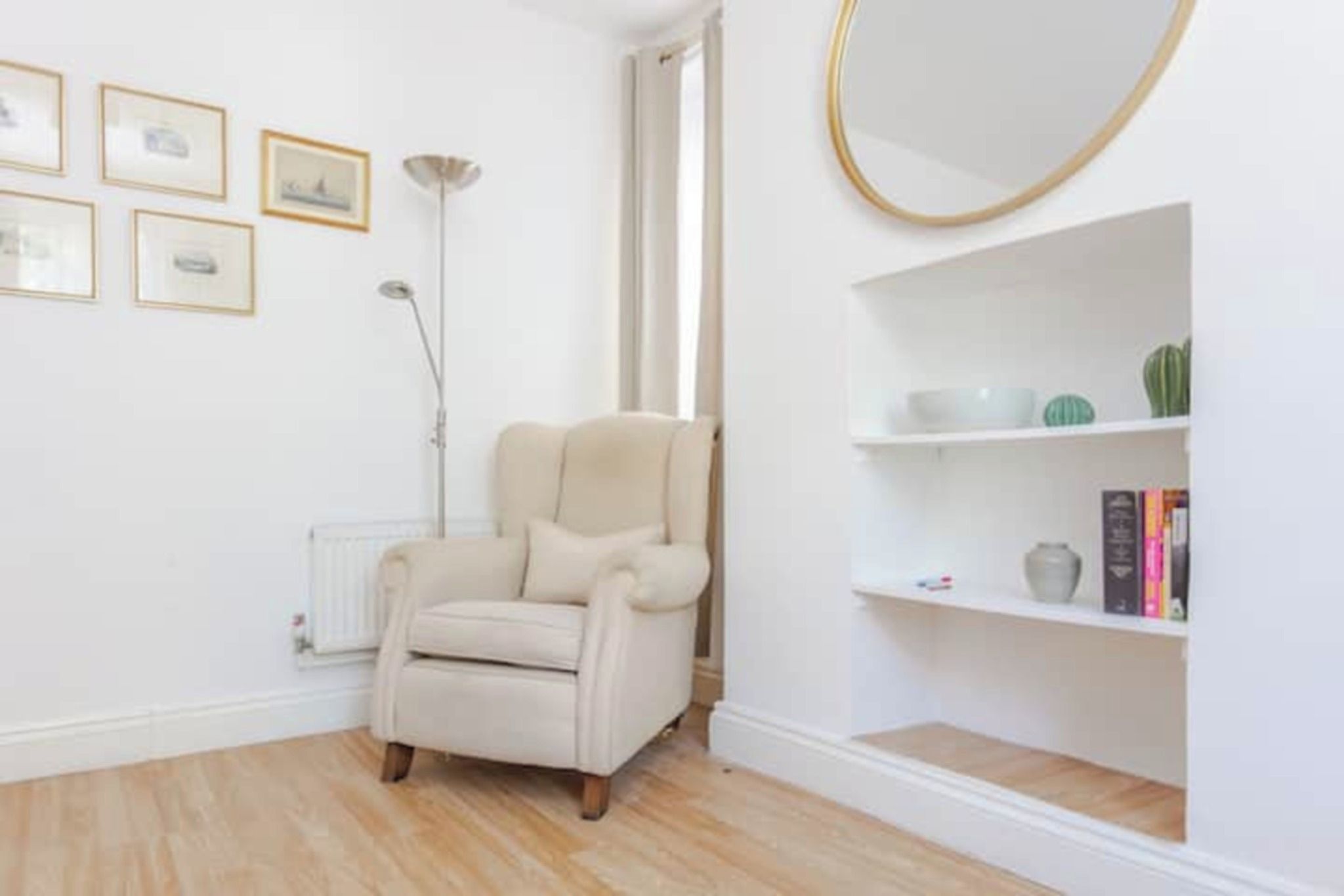 Une pièce comprenant un fauteuil beige, un radiateur, un lampadaire, un miroir rond et une bibliothèque agrémentée d’objets décoratifs.