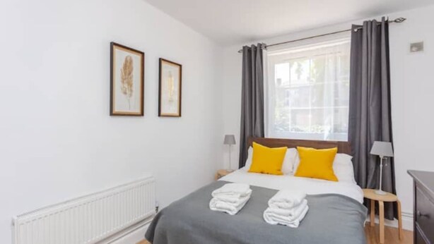 Une chambre d’hôtel avec un lit, deux tableaux encadrés au mur, une fenêtre avec des rideaux et un radiateur.