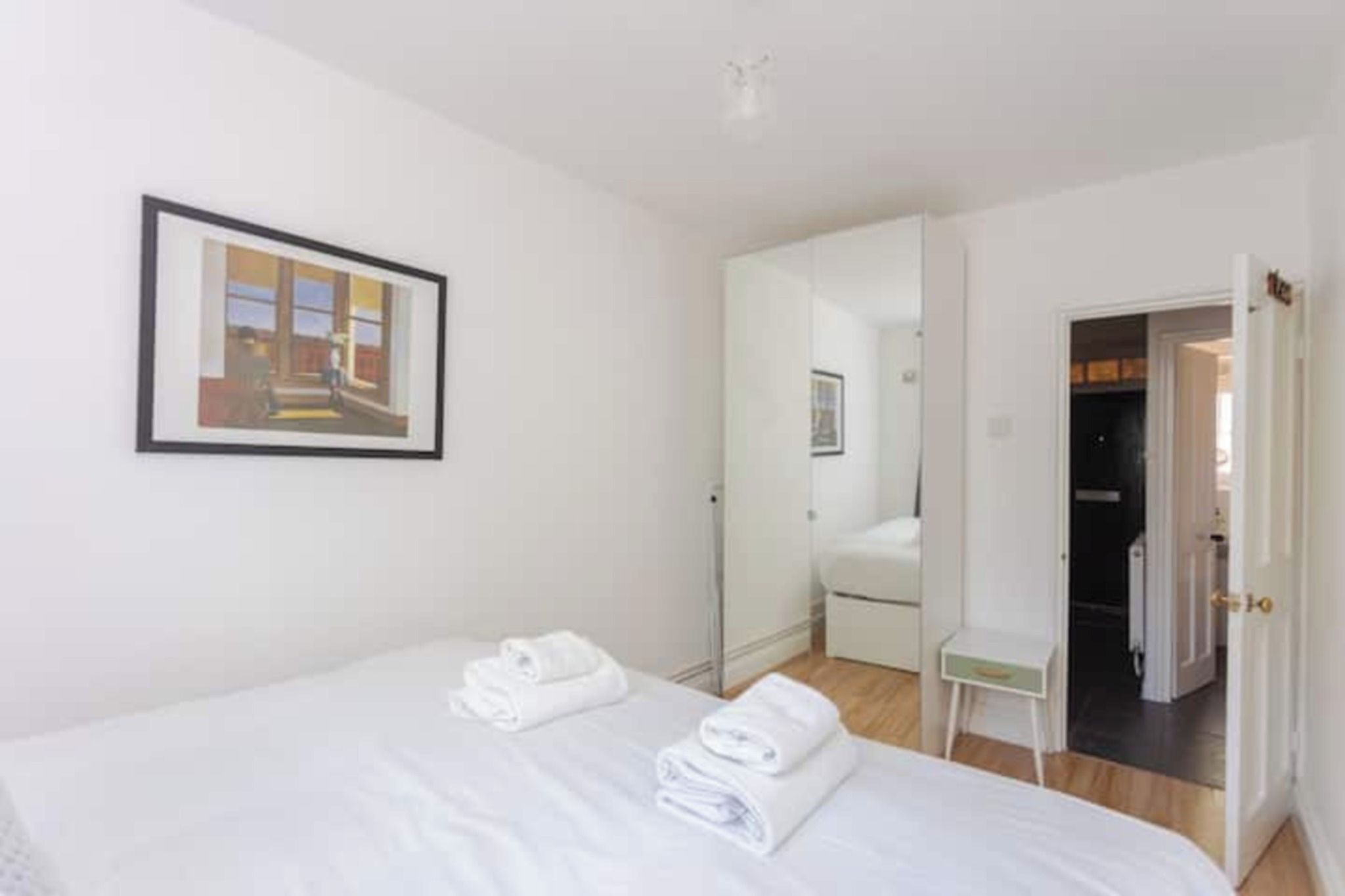 A hotel room with a bed, a framed picture on the wall, a bathroom visible through an open door, and a small bedside table.