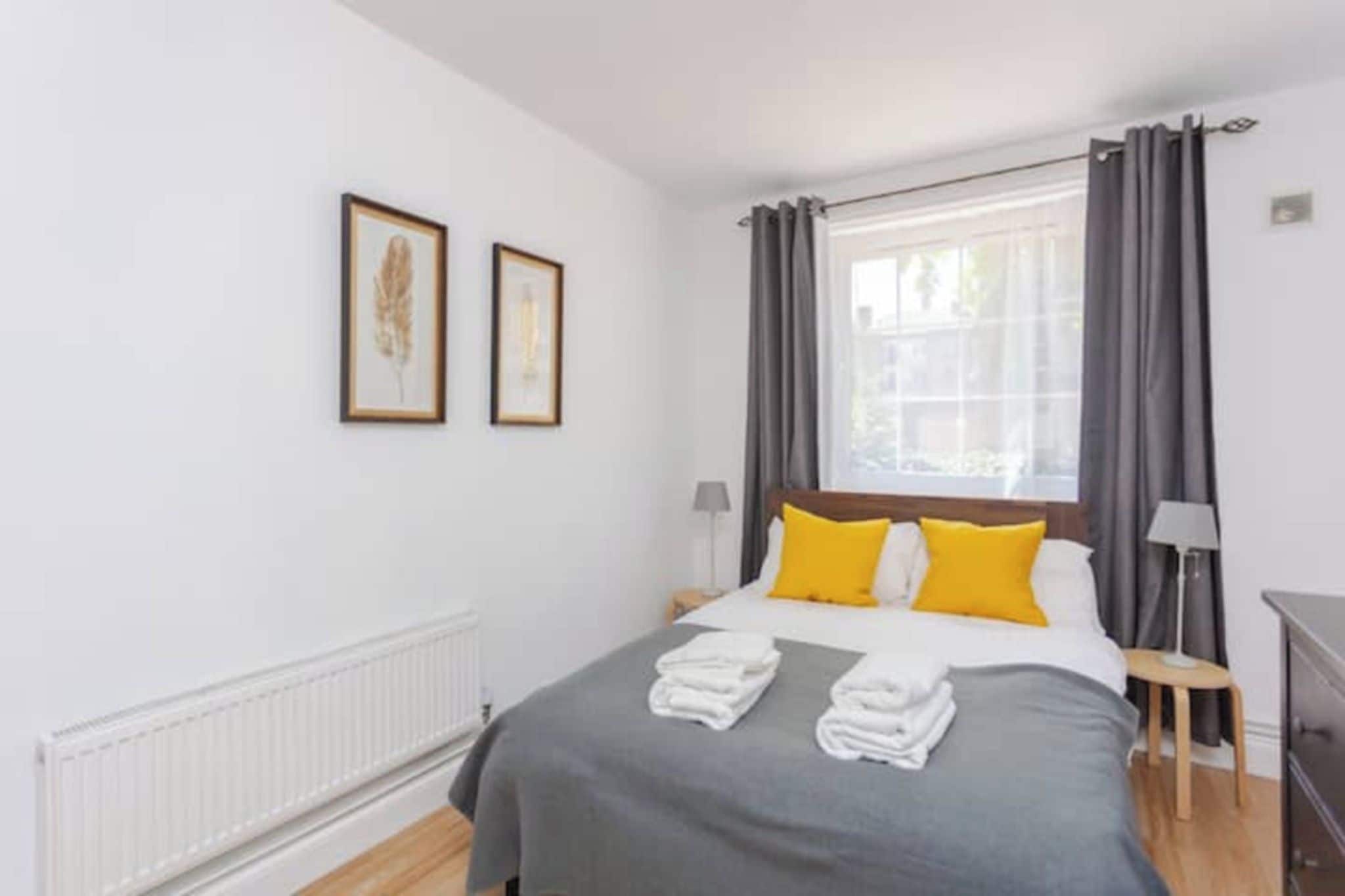 A hotel room with a bed, two framed pictures, a window with curtains, a radiator, and a lamp.