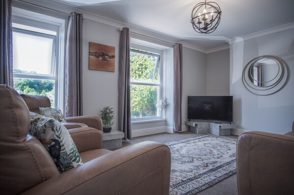 Een woonkamer met leren banken, een flatscreen televisie, een groot raam met gordijnen, een kroonluchter en een ronde spiegel aan de muur.