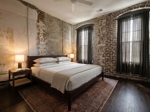 Un dormitorio con pared de ladrillo, una cama grande con ropa de cama blanca, dos lámparas de noche y una ventana con cortinas.