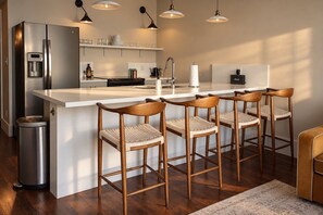 Una cocina moderna con refrigerador de acero inoxidable, dispensador de agua integrado, cafetera y barra con taburetes de madera.