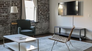 A modern living room with a brick wall, a flat-screen TV, a green armchair, a white coffee table, and a wicker chair. - The Grant by Black Swan (Savannah)