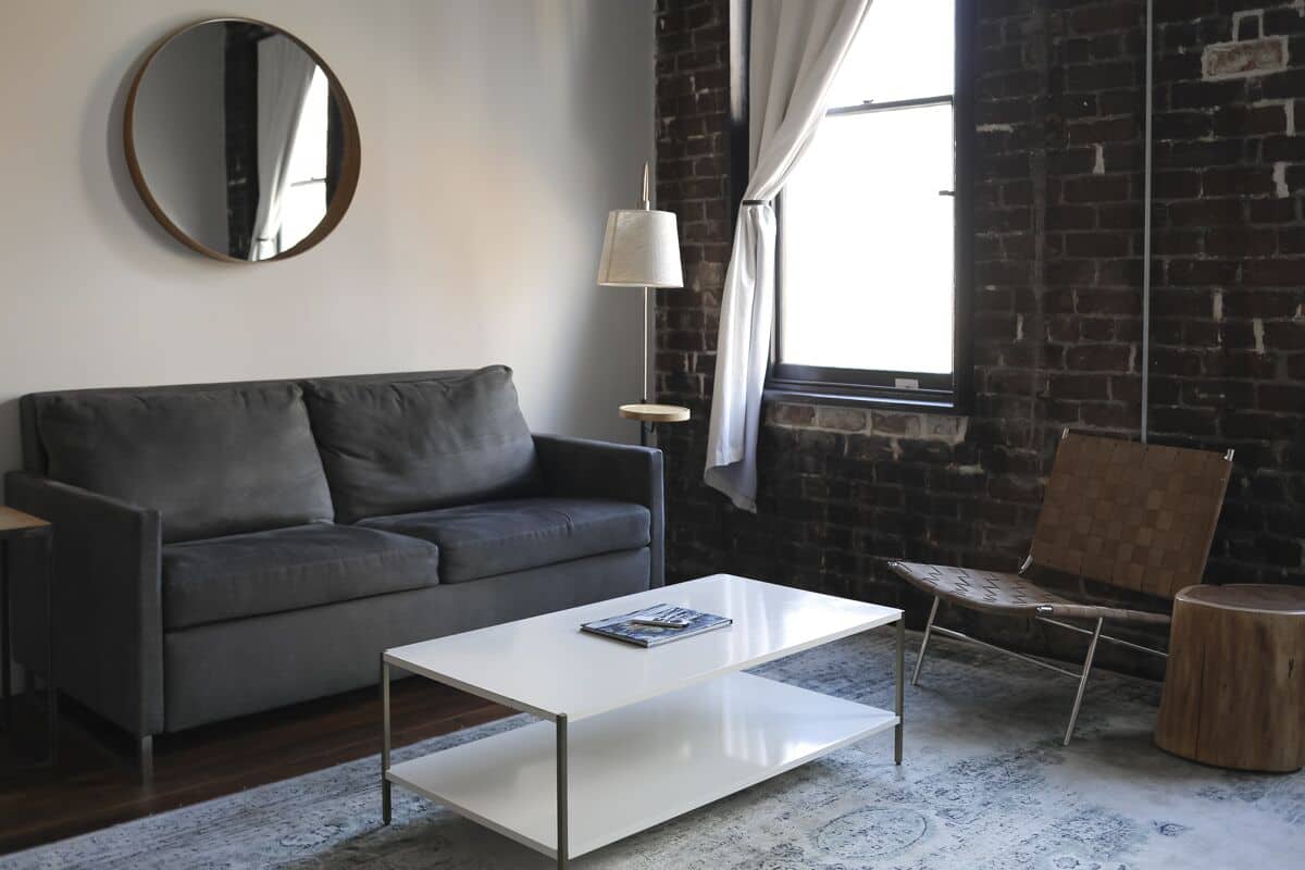 A modern living room with a grey sofa, a white coffee table, a round mirror, a floor lamp, and a brick wall.